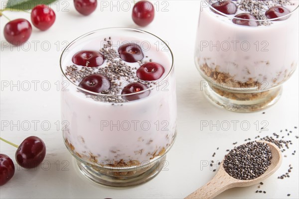 Yoghurt with cherries, chia seeds and granola in glass with wooden spoon on white wooden background
