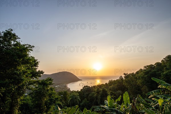 View from a mountain onto a secluded bay with a sandy beach and mangrove forest. The sun sets over