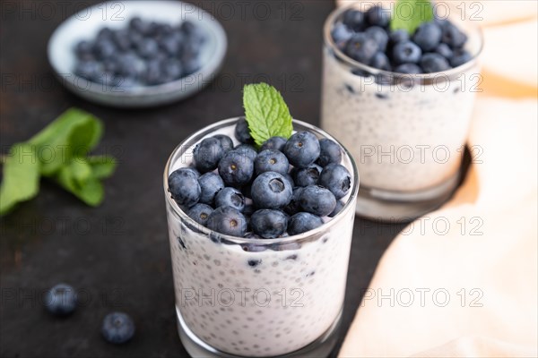 Yogurt with blueberry and chia in glass on black concrete background and orange linen textile. Side