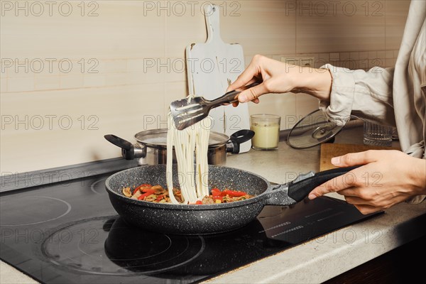 Unrecognizable woman transfers noodles with kitchen tongs into a frying pan with vegetable dressing