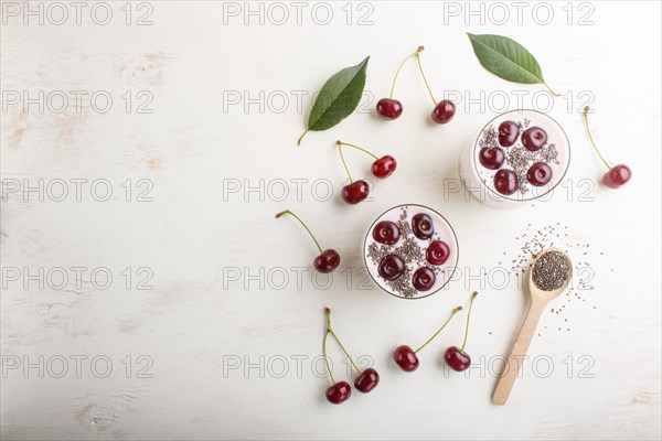 Yoghurt with cherries, chia seeds and granola in glass with wooden spoon on white wooden background