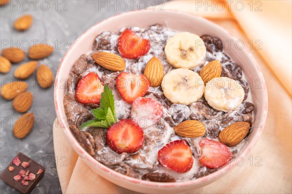 Chocolate cornflakes with milk, strawberry and almonds in ceramic bowl on gray concrete background