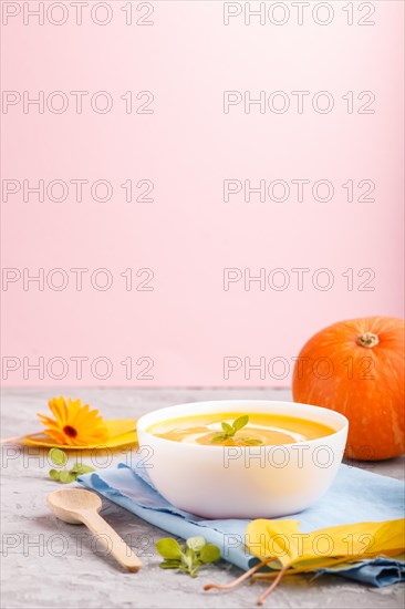 Traditional pumpkin cream soup with in white bowl on a gray and pink background with blue napkin.