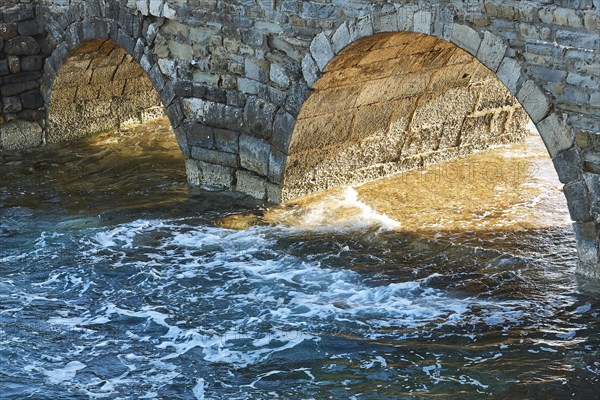 Sunlight shines through stone arches of an old bridge with foaming sea waves below, sea fortress