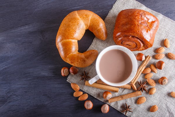 A cup of hot chocolate with nuts, buns and spices on linen napkin and black wooden background. copy