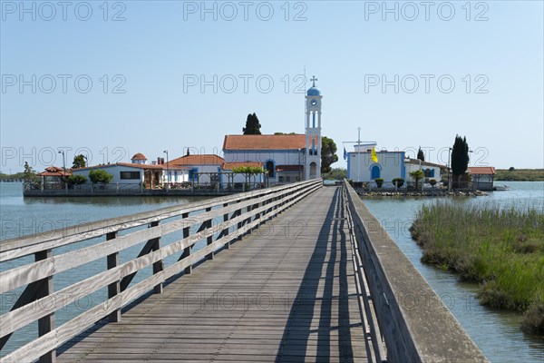 A wooden footbridge leads over calm waters to a monastery under a clear blue sky, Monastery of St
