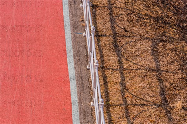 Looking down onto red bike path next to metal guardrail casting its shadow on brown grassy area in