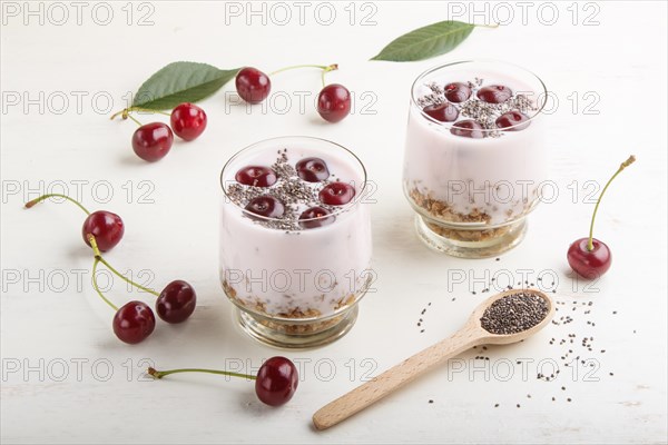 Yoghurt with cherries, chia seeds and granola in glass with wooden spoon on white wooden background