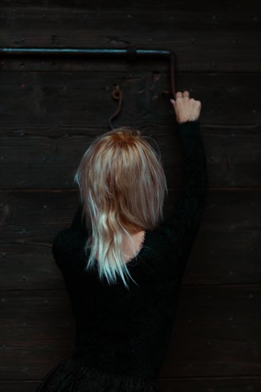 Back view of a blonde thin white woman dressed in black reaching up to a rusty antique metal lock