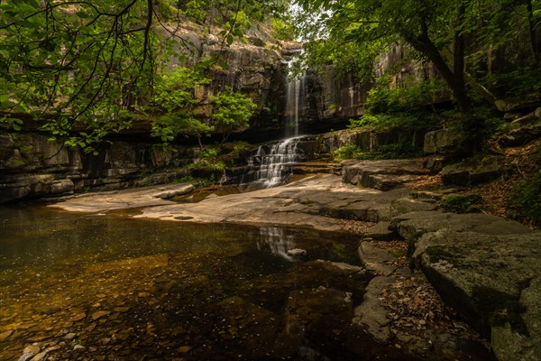 A secluded waterfall spills over ancient rocks into a still pool, shaded by a wooded canopy, in