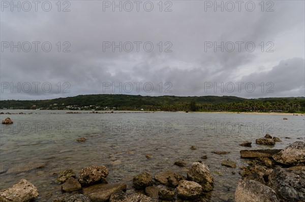 Seascape of rocky shoreline on a cloudy day with buildings on tree lined shore in the distance in