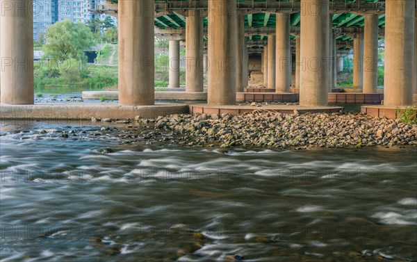 Flowing river under an overpass with concrete columns and greenery around, in South Korea