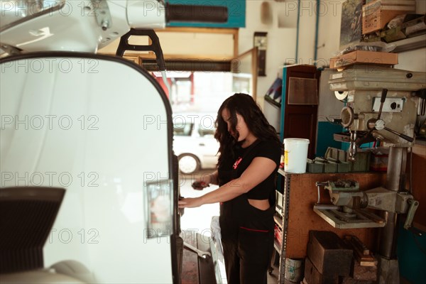 Mechanic standing by a classic scooter in a sunlit workshop full of tools, latino female in