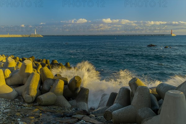 Waves crashing against tetrapods laying on ocean shoreline with two lighthouses on piers on horizon