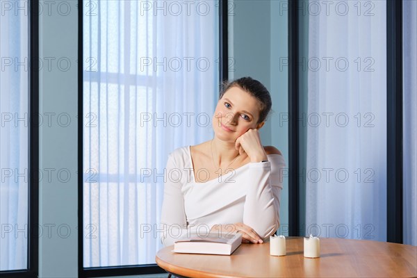 Dreamy woman sits at a wooden table with a book, props her head up with her fist and looks with a