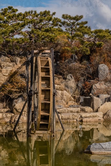 Close-up of a water wheel with its reflection in a pond, set against a rocky backdrop, in South