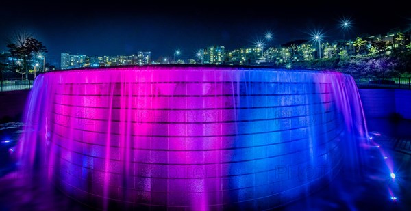 Night scene of waterfall lit up with blue and red lights with the lights of buildings of a town in