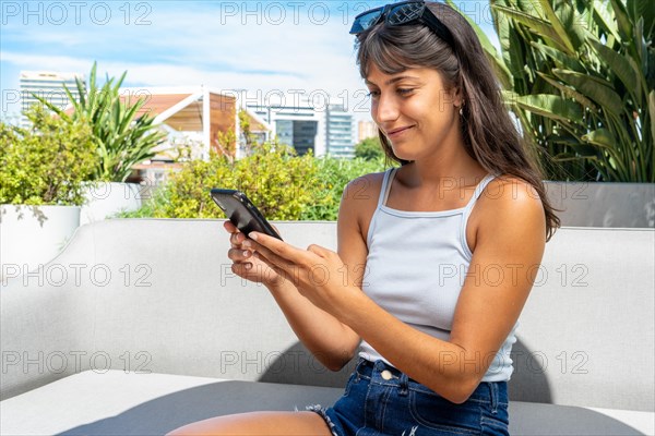 A woman is sitting on a couch and looking at her cell phone. She is wearing a white tank top and