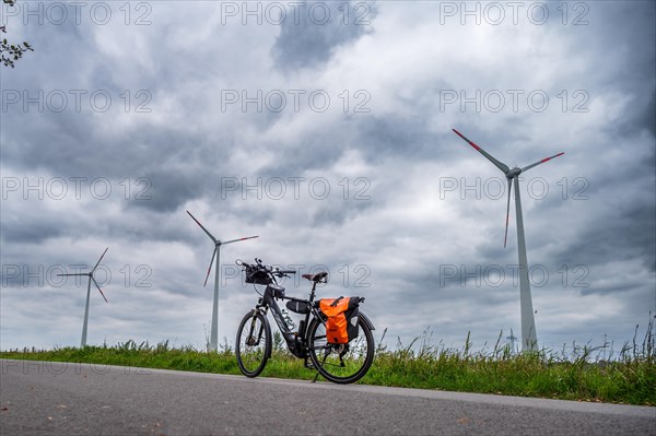 A parked bicycle at the roadside with wind turbines in the background under a cloudy sky, Leer,