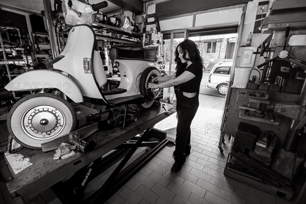 Woman mechanic working on repairing a white moped italian vintage scooter, surrounded by tools in a