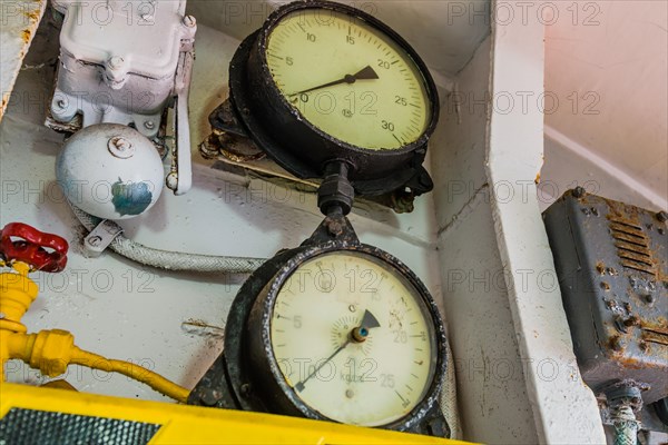 Old pressure gauges mounted above yellow and black striped ledge with valves and bell in background