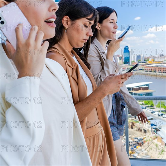 Three women are standing on a balcony, each holding a cell phone. They are all dressed in business