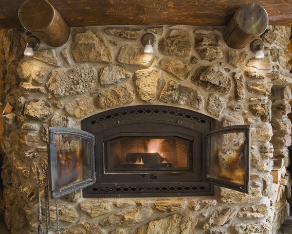 Lit fieldstone and porous rock fireplace in living room inside handcrafted red cedar log cabin