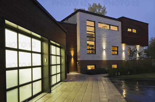 Illuminated two car garage and beige stone with brown cedar wood siding modern cubist style home