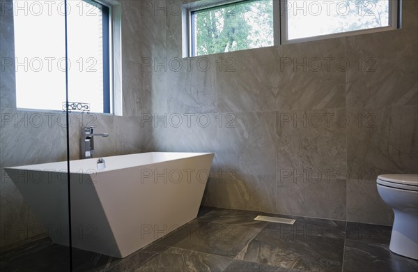 White freestanding vessel shaped bathtub in bathroom with grey ceramic tile floor and walls on