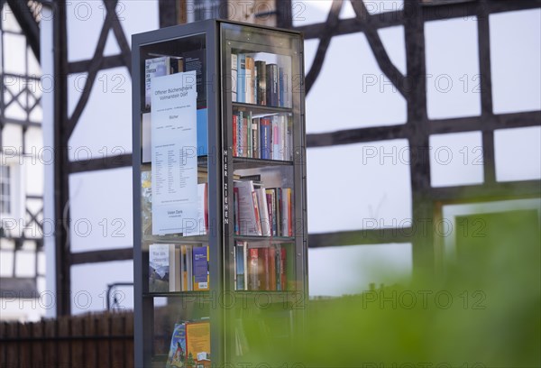Open bookcase in front of half-timbered house and fence, many colourful books in a glass display