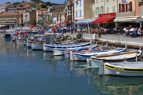Cassis, the harbour, people strolling along a promenade with boats and pastel-coloured houses,