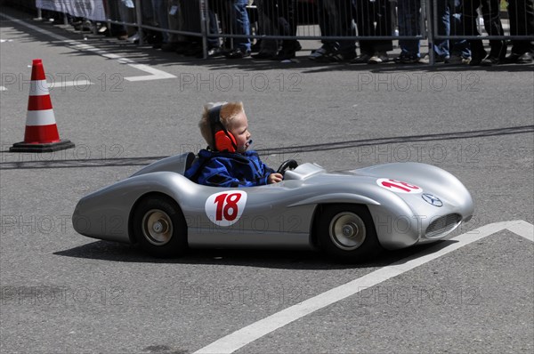 Child driving a soapbox during a competition, watched by spectators at the side of the track,