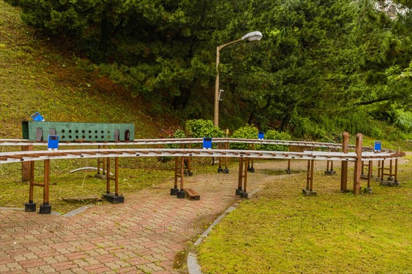 Miniature outdoor train station with benches and lampposts surrounded by trees on a cloudy day, in