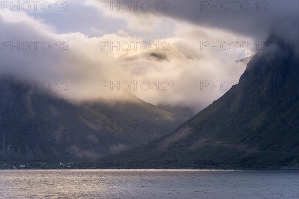 View over the Flakstadpollen to mountains in clouds with incoming sunlight. The small village of