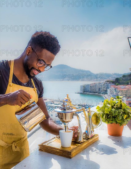 Happy person making coffee outdoors with a scenic coastal view in bright daylight, Vertical aspect