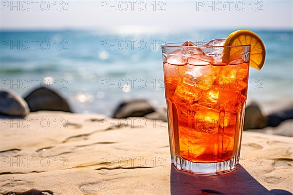 Glass with orange cocktail drink with ice cubes and orange fruit slice with beach in background. KI