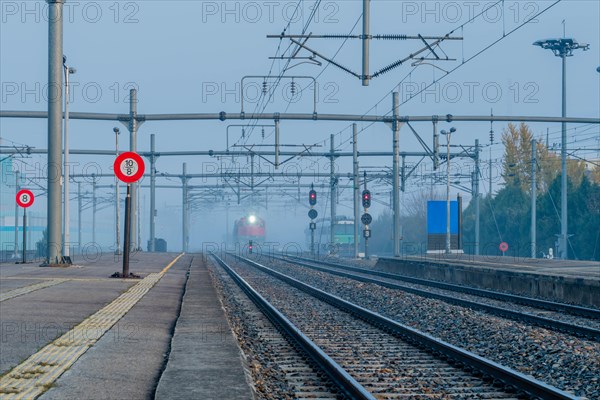 Desolate railway station in foggy conditions at dawn with signal lights on and a train approaching,