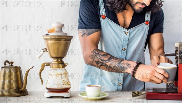 Bearded hipster man with tattoos using a manual coffee grinder next to a vintage kettle,