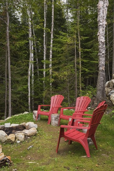 Four bright red plastic Adirondack chairs around rock edged firepit dug into the ground in backyard