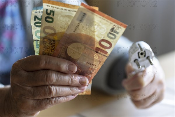 Senior citizen holding money and a power cable with plug in her hand at home, symbolising energy
