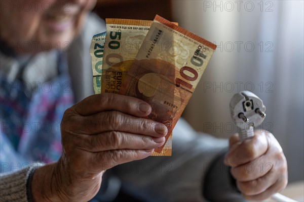 Senior citizen holding money and a power cable with plug in her hand at home, symbolising energy