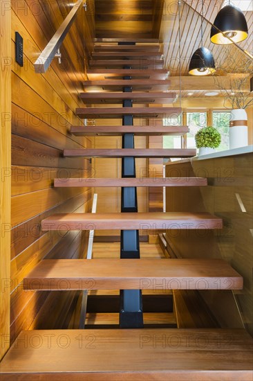 Looking up illuminated wooden staircase with opened steps and clear glass railing leading to