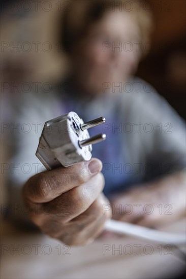 Senior citizen holding a power cable with plug in her hand at home, symbolising energy costs and