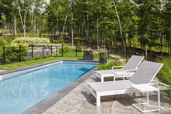 Two white long chairs on edge of in-ground swimming pool enclosed by clear glass and black metal