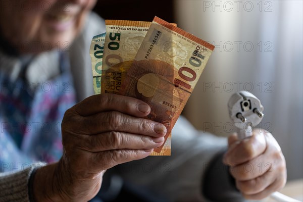 Senior citizen holding money and a power cable with plug in her hand at home, symbolising energy