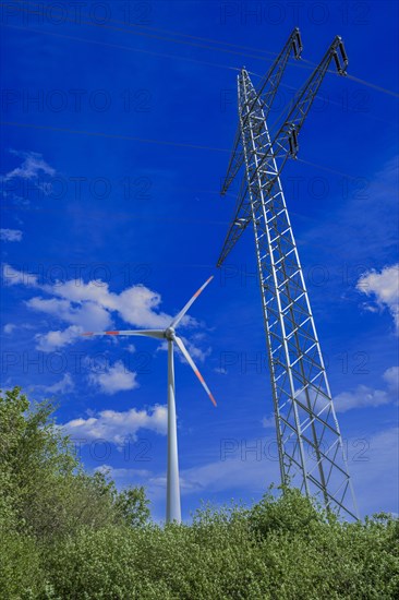 Power pylon with high-voltage lines and wind turbine behind green trees at the Avacon substation