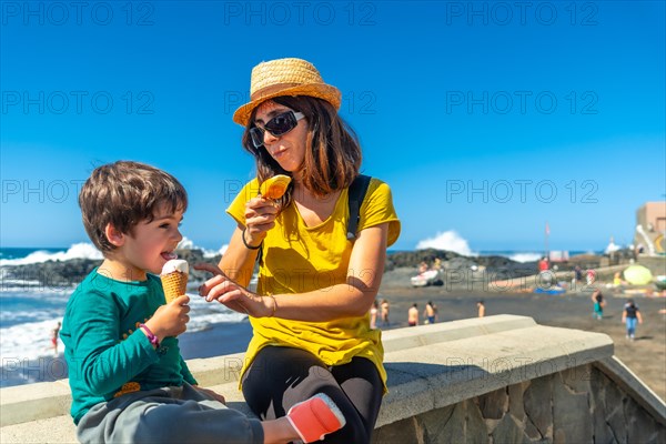 A woman and a child are sitting on a wall near the ocean, eating ice cream. The woman is wearing a