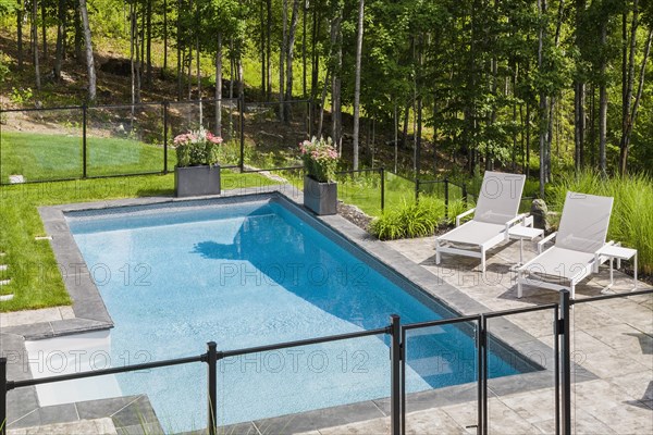 Two white long chairs on edge of in-ground swimming pool enclosed by clear glass and black metal