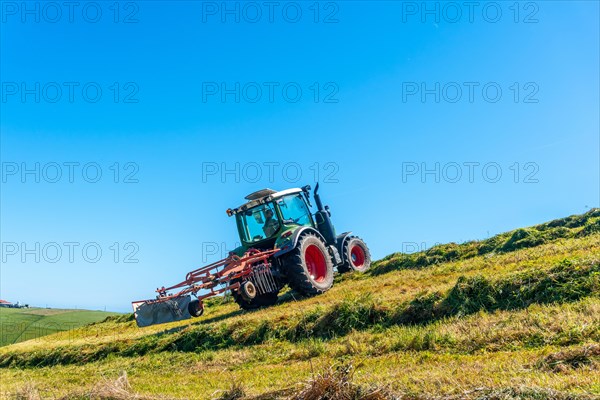 A tractor is driving down a hillside, plowing the grass. The sky is clear and blue, and the sun is