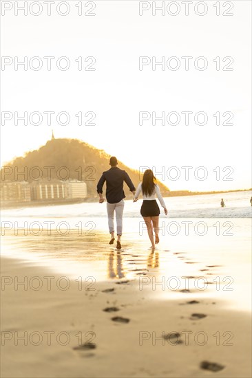 Vertical rear view of a tender young couple walking holding hands on the edge of the sea water